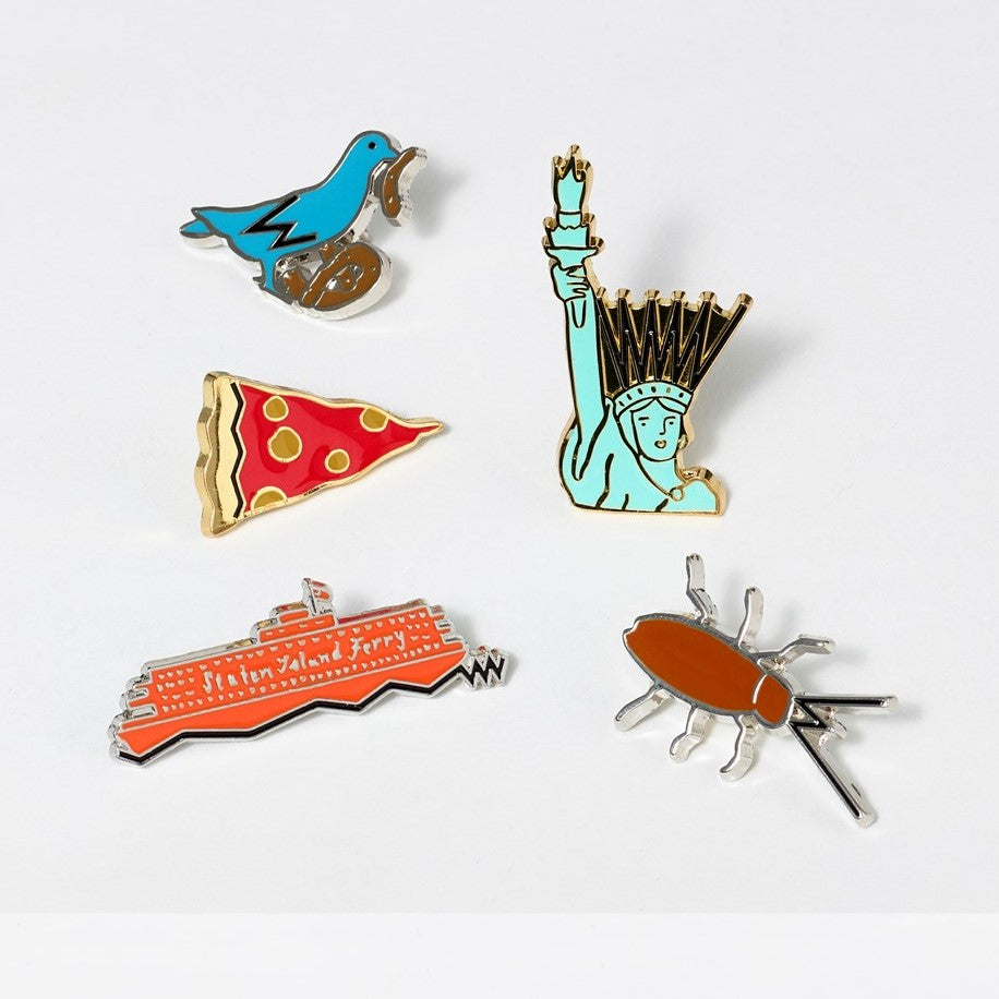 Assortment of hard enamel and metal pins illustrated by Tamara Shopsin, featuring iconic New York landmarks; Pigeon, Lady Liberty, Roach, Staten Island Ferry, and Pizza slice.