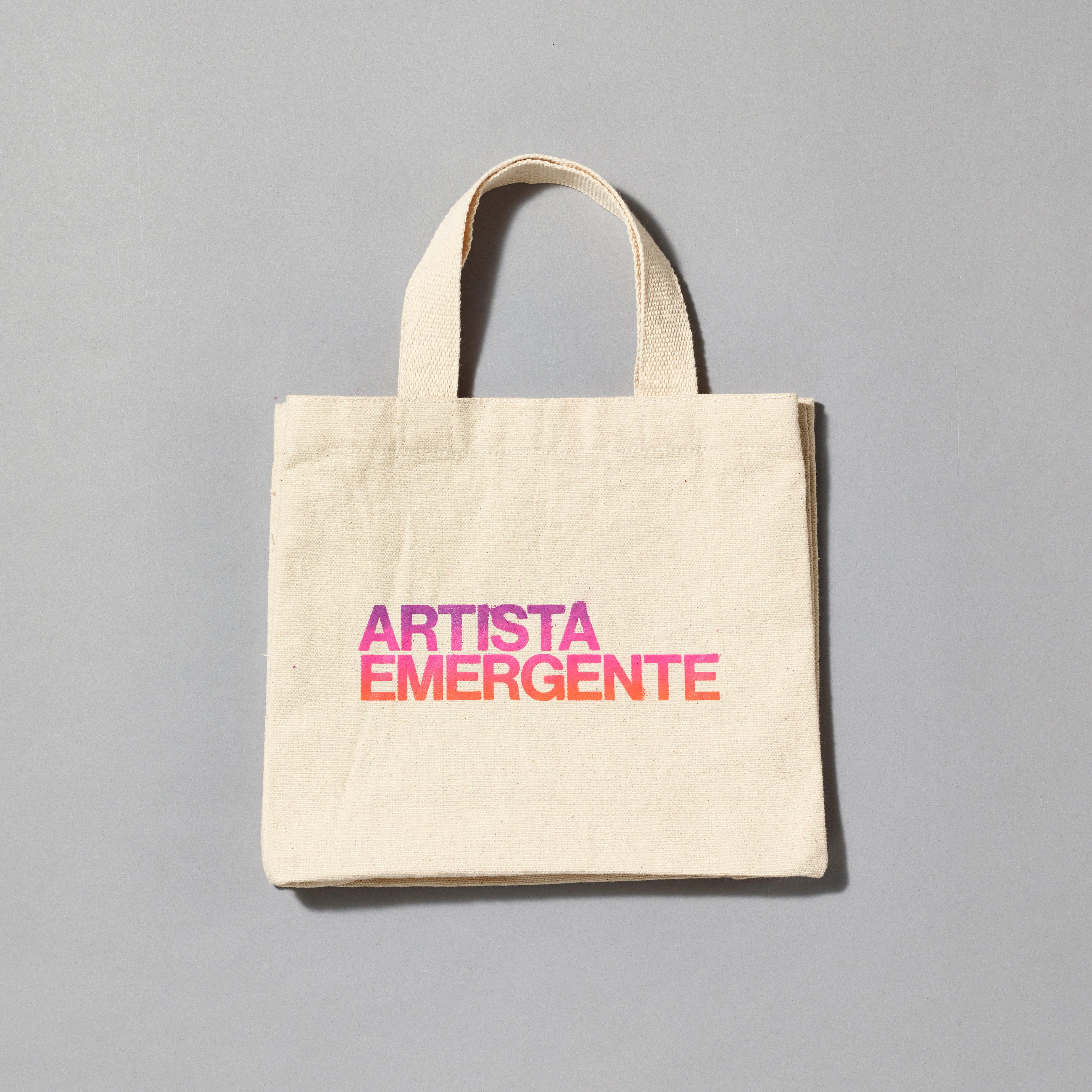 100% cotton canvas tote with Artista Emergente printed in a purple, pink, and orange gradient. Measures 8.75" H x 10.75" L x 3" W