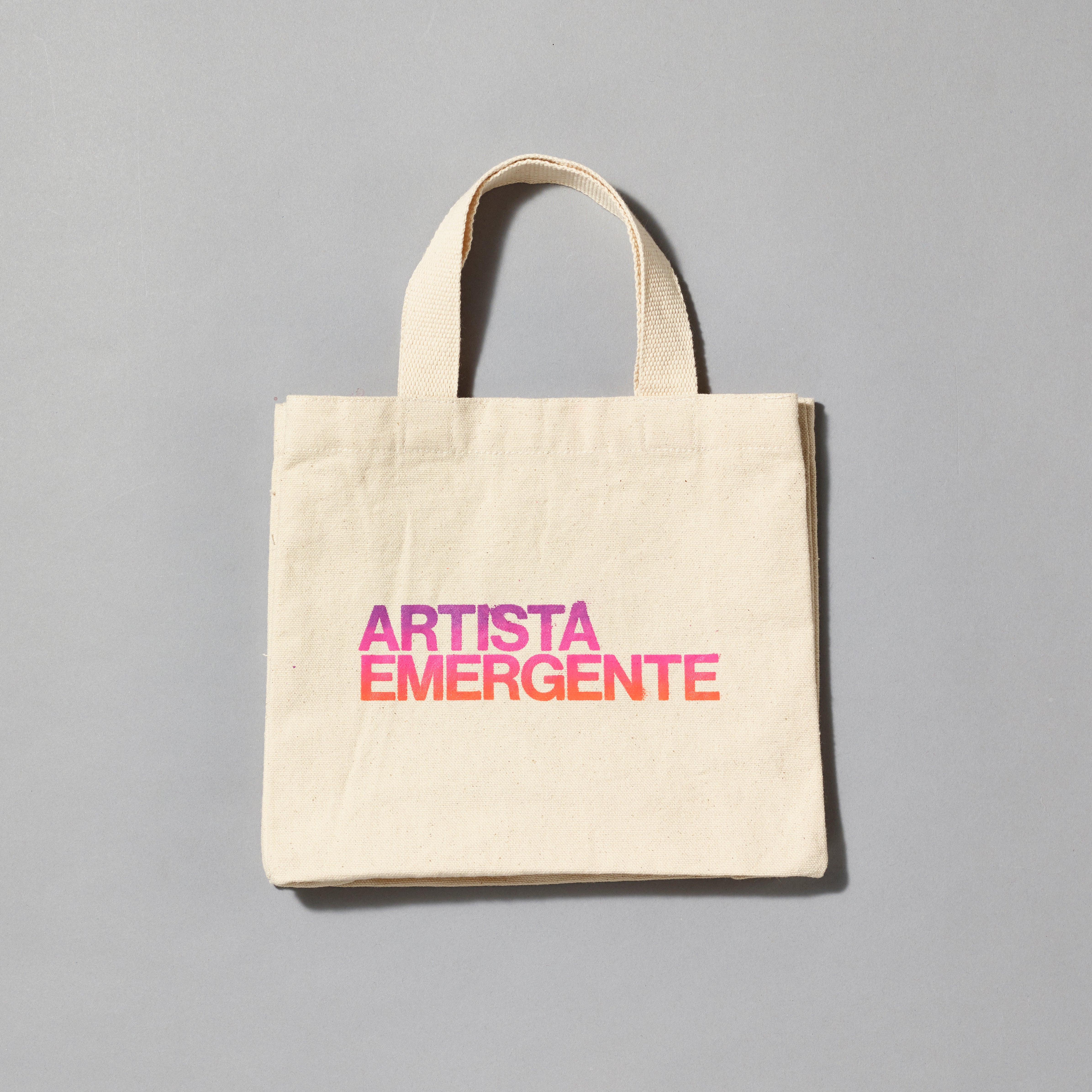 100% cotton canvas tote with Artista Emergente printed in a purple, pink, and orange gradient. Measures 8.75" H x 10.75" L x 3" W