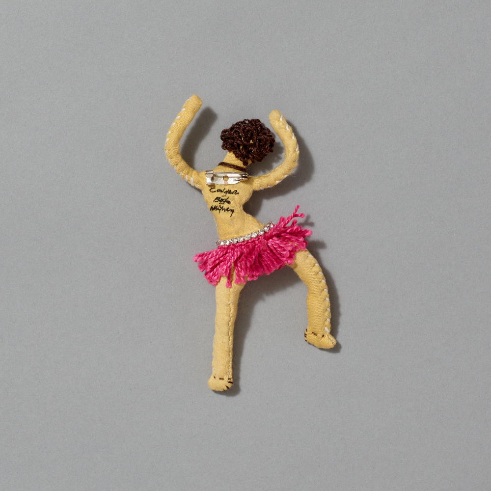 Textile brooch of a dancer with pink skirt and yellow body on gray background