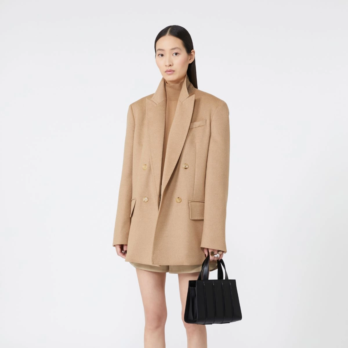 Model wearing a beige coat holding a black handbag on a white background