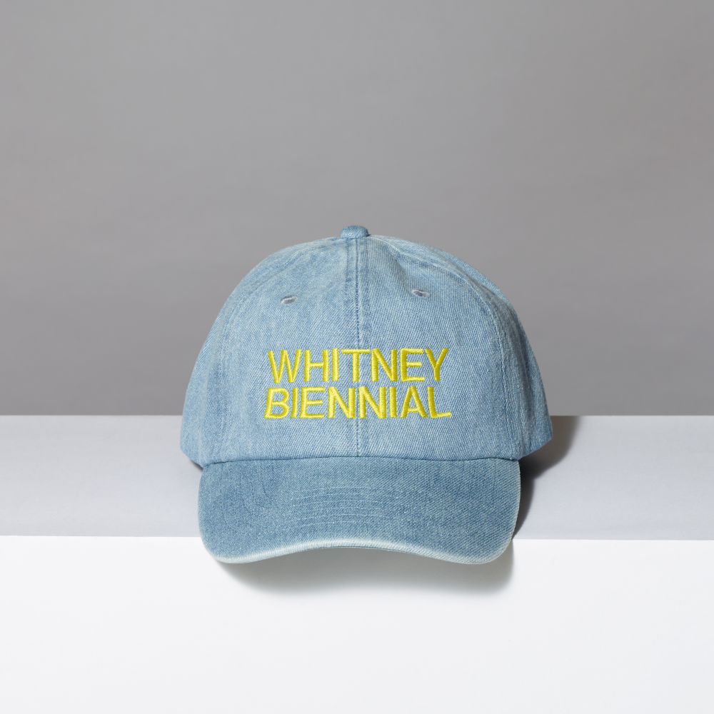 Denim cap with 'WHITNEY BIENNIAL' text on a gray background