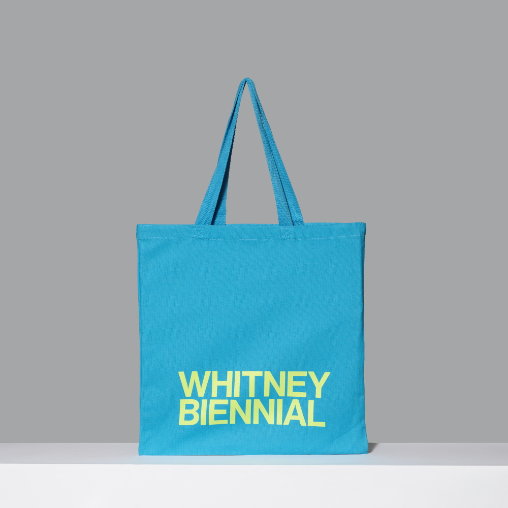 Blue tote bag with 'WHITNEY BIENNIAL' text on a gray background