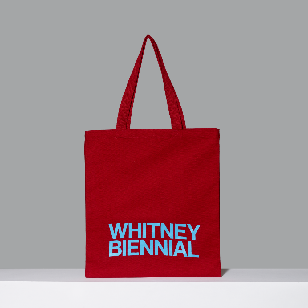 Red tote bag with 'WHITNEY BIENNIAL' text on a gray background