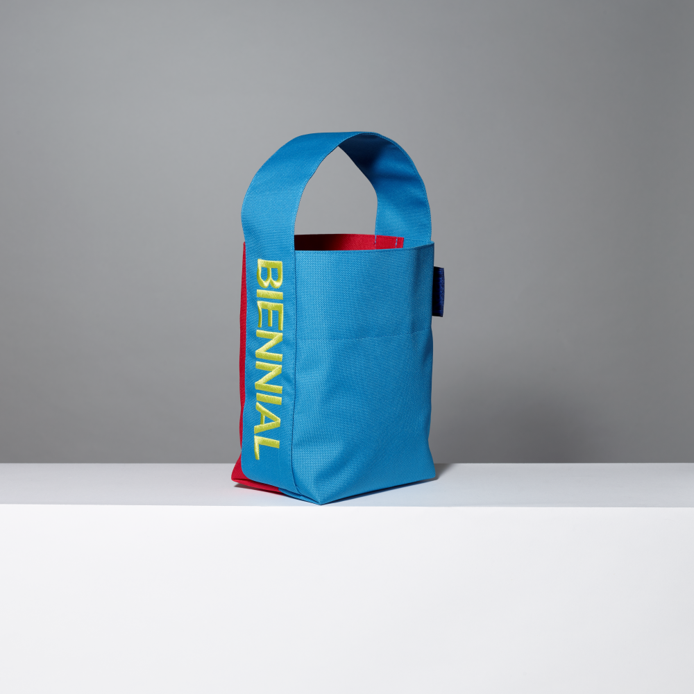 Blue bag with red interior and 'BIENNIAL' yellow text on a gray background