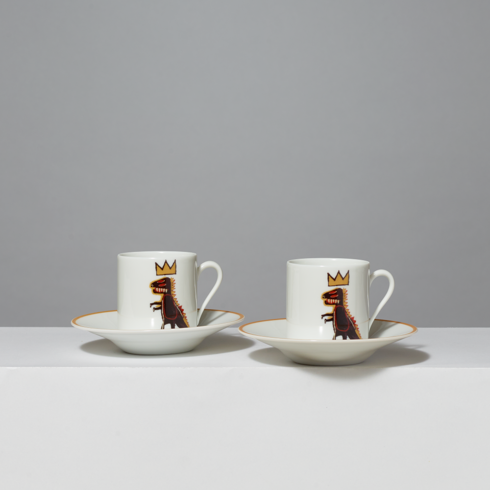 Two white ceramic espresso cups with saucers, each decorated with Basquiat's Pez Dispenser dinosaur wearing a gold crown, set against a neutral gray background.