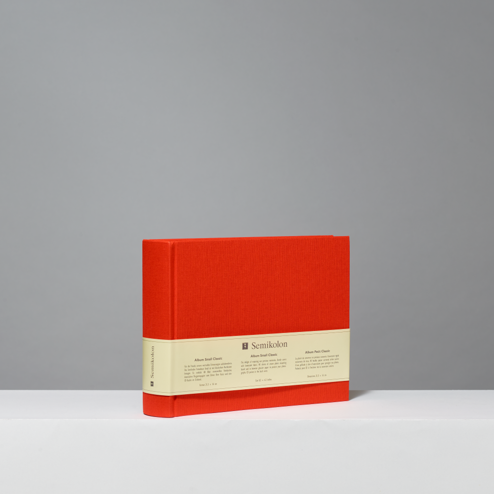 An orange hardcover photo album standing upright on a white surface against a gray background. A beige paper band wraps around the middle, with the brand name 'Semikolon' printed on it.