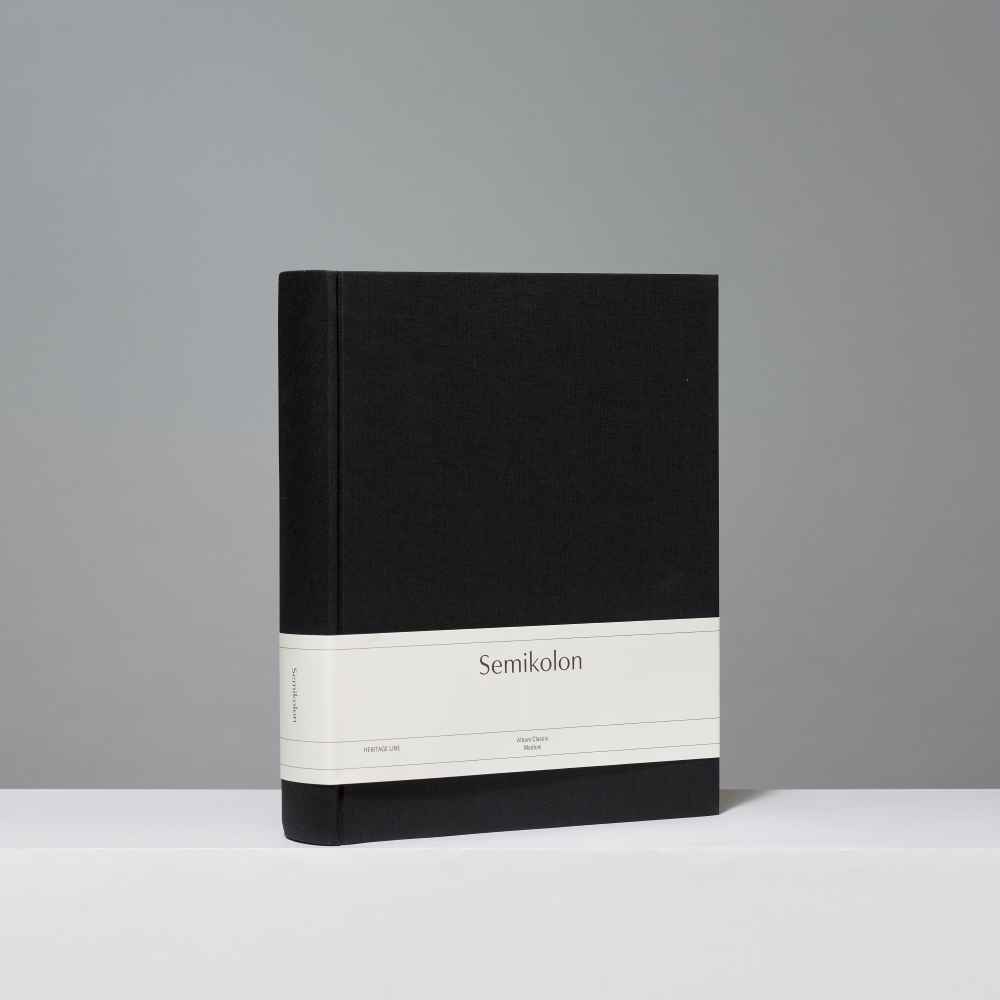 A hardcover photo album with a black textured cover and a white paper band around the middle, standing upright on a white surface against a gray background.