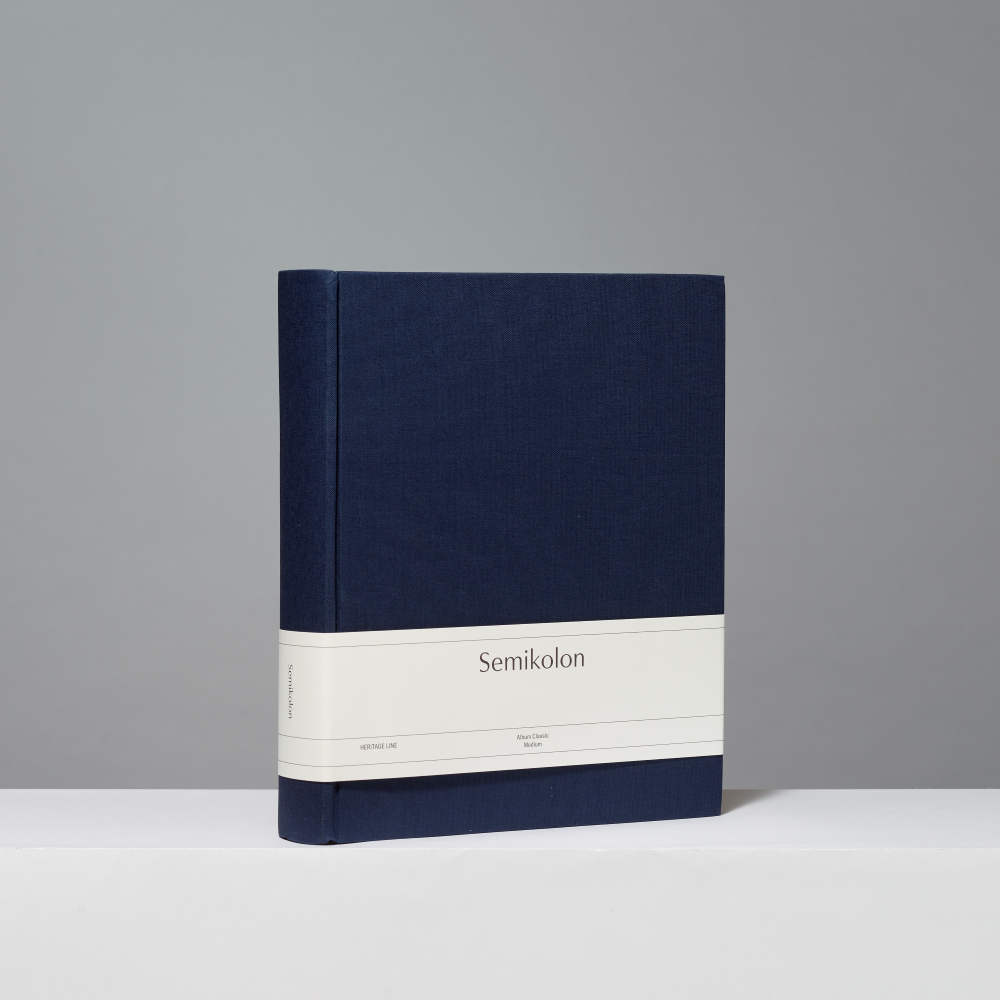 A hardcover photo album standing upright on a white surface against a light gray background. The cover is dark navy blue with a wide white horizontal band around the middle printed with the word "Semikolon.