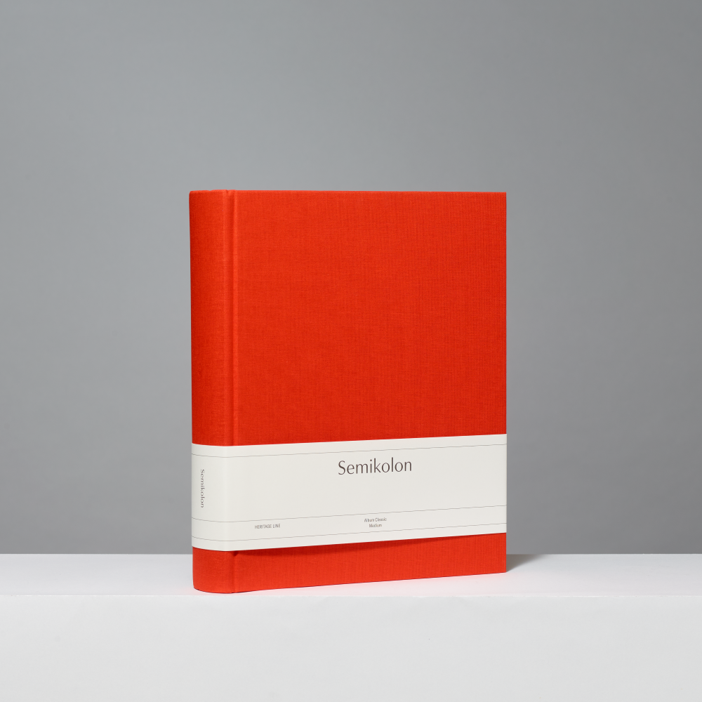A hardcover photo album with a textured orange-red cover, a white paper band around the middle, and the word 'Semikolon' printed on it, photographed against a neutral gray background.