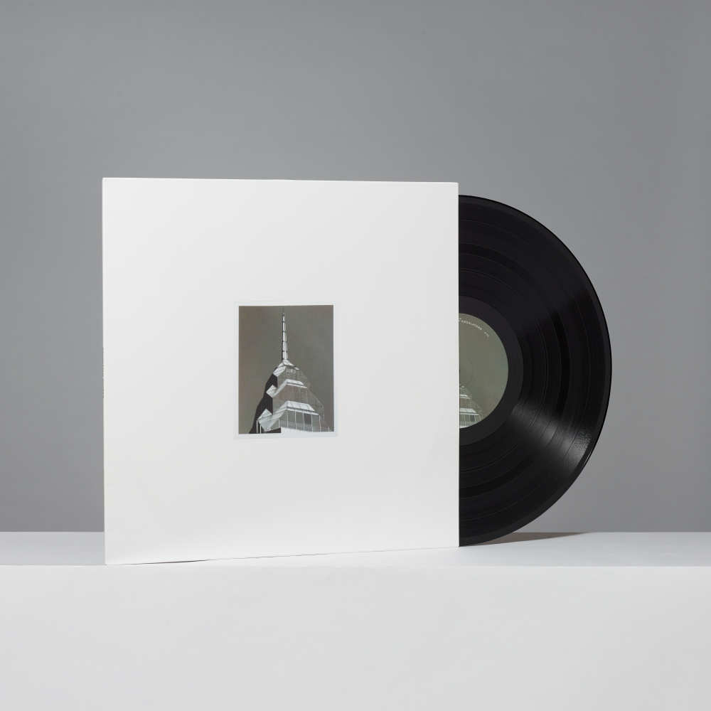 A vinyl record partially pulled from a white square album sleeve, with a small black-and-white architectural photo printed in the center of the sleeve, set against a plain gray background.
