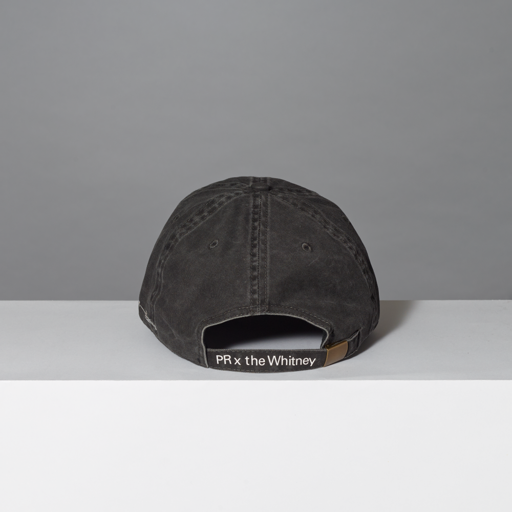 A washed black baseball cap shown from the back, resting on a white surface against a gray background. The back strap features a black patch with white text reading 'PR x the Whitney'.