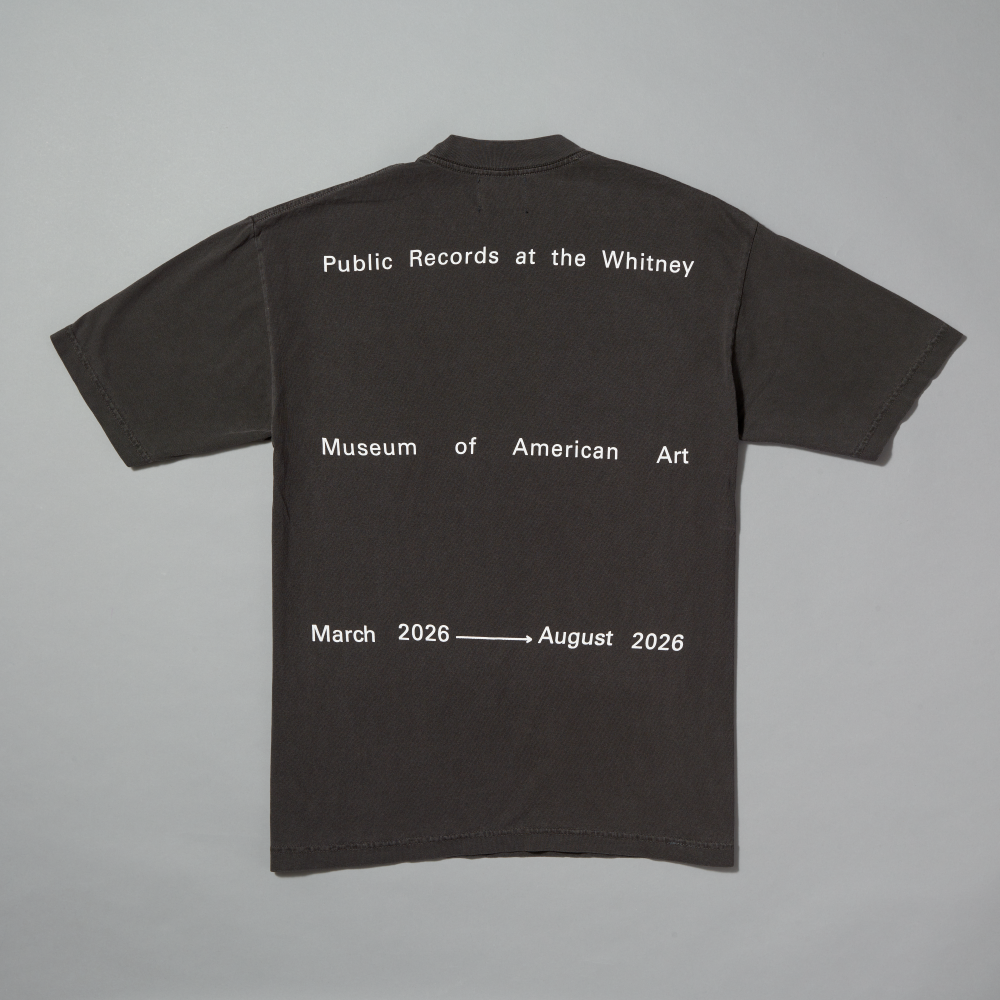 A dark gray short-sleeve t-shirt shown from the back, featuring white text that reads 'Public Records at the Whitney', 'Museum of American Art', and 'March 2026 to August 2026' on a light gray background.