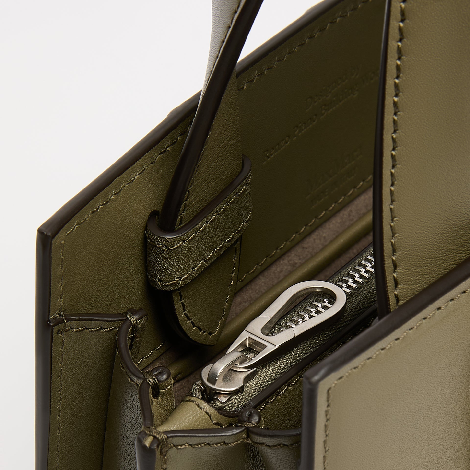 Close-up detail of an olive green leather handbag showing the interior lining, a silver-tone zipper pull, and stitched edges against a light neutral background.