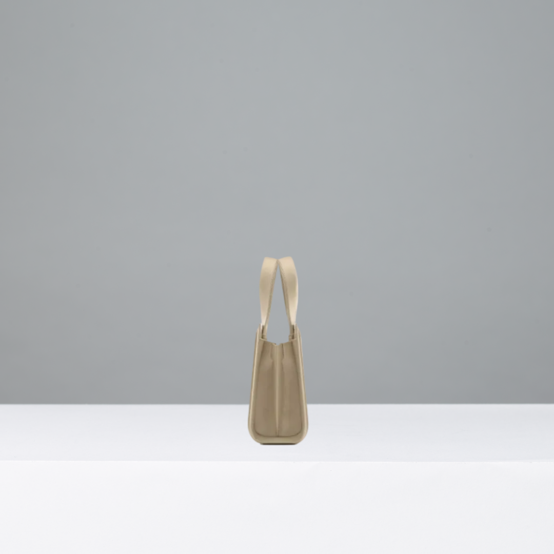 A minimalist taupe leather shoulder bag with a single strap, standing upright on a white surface against a neutral gray background.