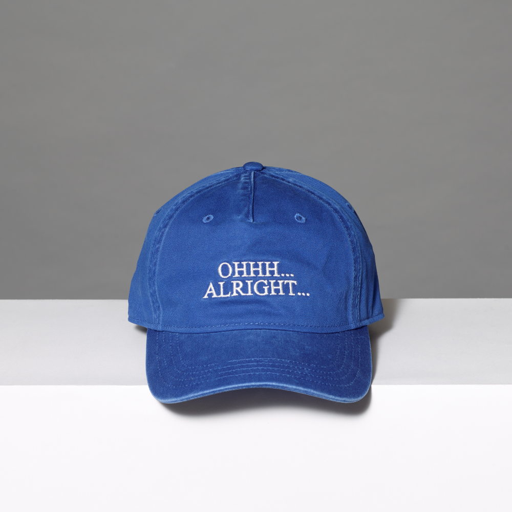 A blue distressed baseball cap with white embroidered text reading 'OHHH... ALRIGHT...' inspired by Roy Lichtenstein's work of the same name on a white surface against a gray background