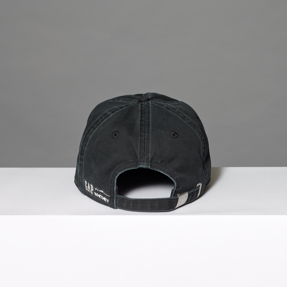 A black cotton baseball cap shown from the back, resting on a white surface against a gray background. The back of the cap features white embroidered text reading 'GAP', 'Lichtenstein' and 'WHITNEY' alongside a silver metal buckle.
