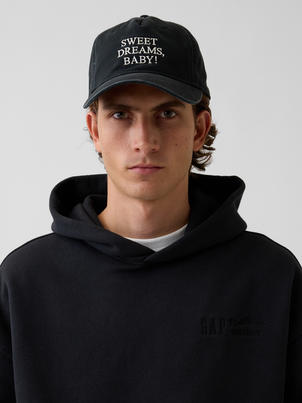 A model wearing a black baseball cap with 'SWEET DREAMS, BABY!' embroidered in white on the front, inspired by Roy Lichtenstein's work of the same name paired with a black hoodie featuring a subtle tonal logo on the left chest, set against a plain light gray background