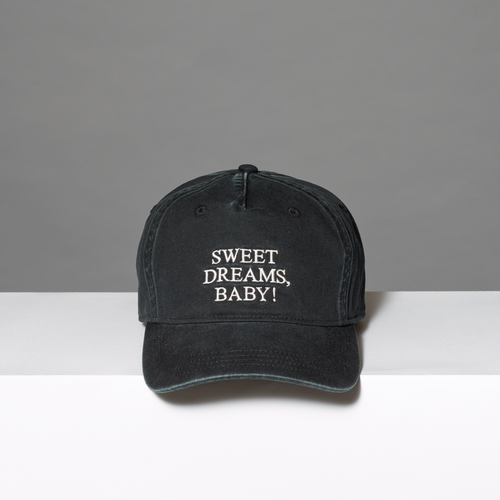 A washed black baseball cap with white embroidered text reading 'SWEET DREAMS, BABY!' inspired by Roy Lichtenstein's work of the same title on the front, displayed on a white surface against a gray background