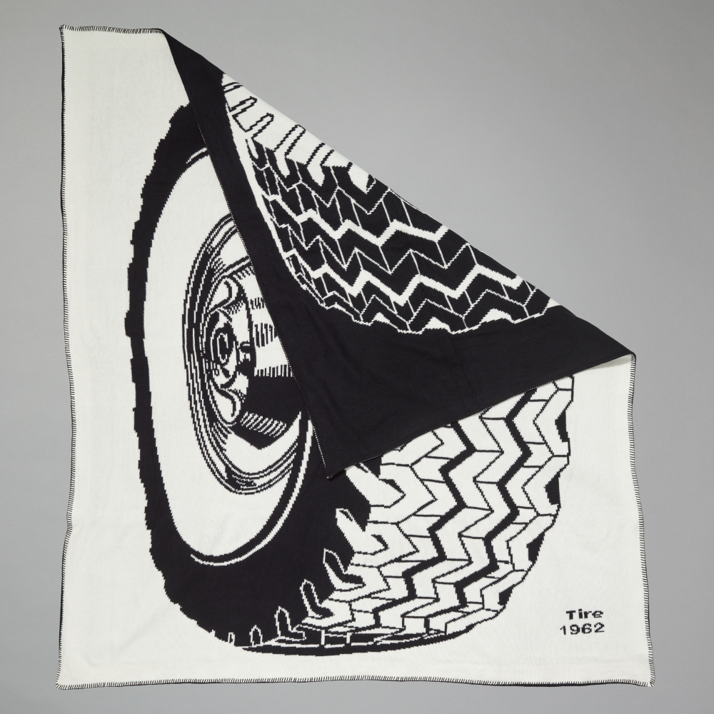 An off-white throw blanket with a large black-and-white illustration of a tire and wheel inspired by Roy Lichtenstein's work Tire folded diagonally on a neutral gray background