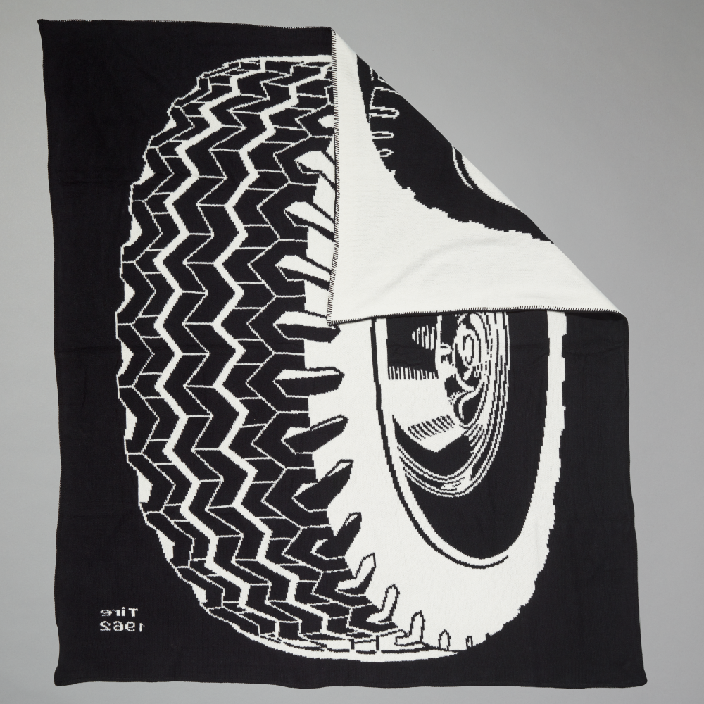 A black throw blanket featuring a bold white graphic print of a tire and wheel inspired by Roy Lichtenstein's Tire, shown with one corner folded over to reveal a white lining, set against a solid gray background