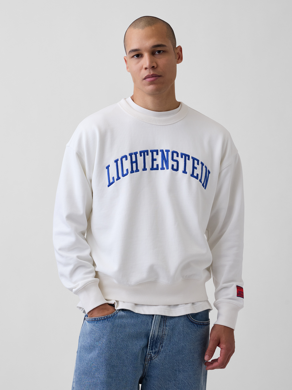 A model wearing a white crewneck sweatshirt with 'LICHTENSTEIN' printed in blue arched letters across the chest, paired with blue jeans, against a plain light gray background