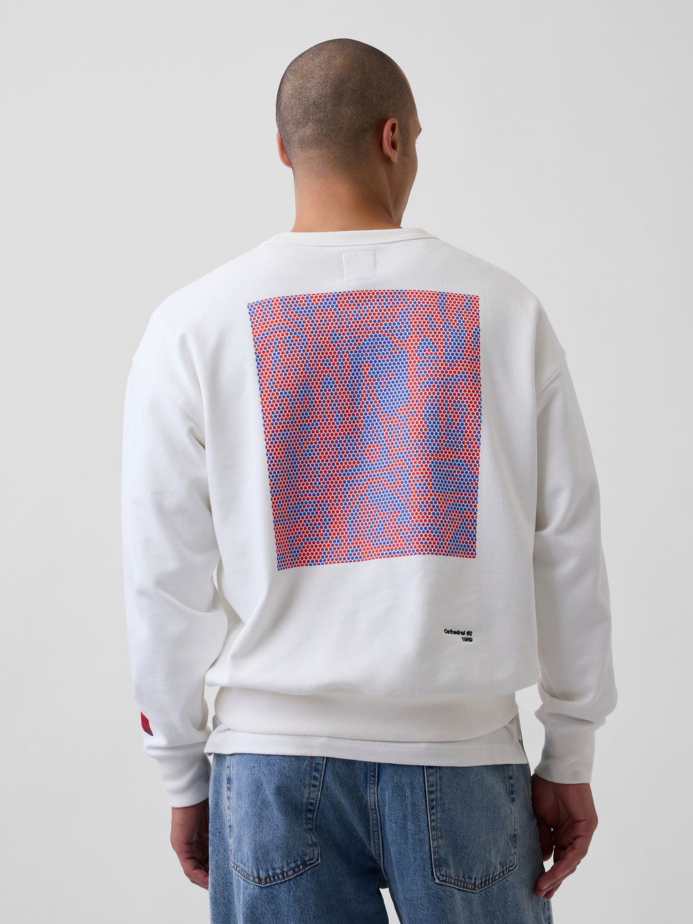 A model viewed from behind wearing a white crewneck sweatshirt with a large red and blue geometric print inspired by Roy Lichtenstein's work Cathedral #2 on the back, paired with blue jeans, against a light gray studio background