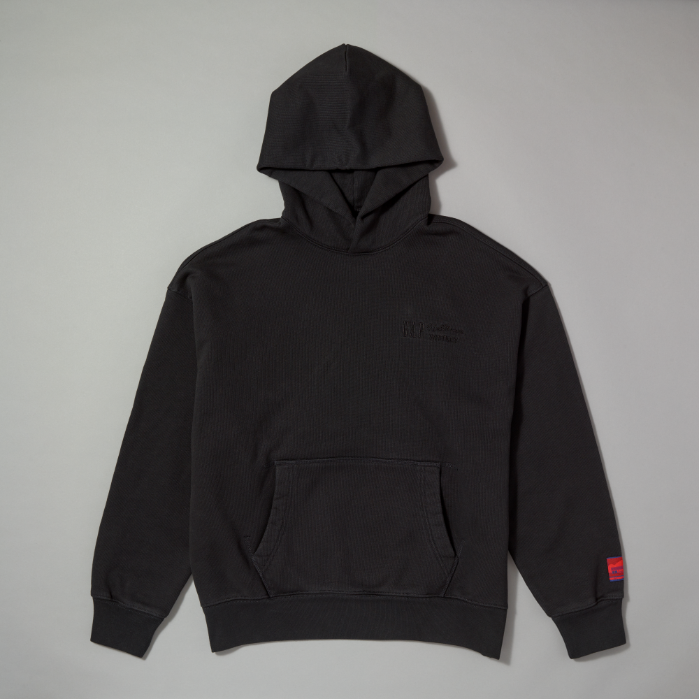 A black hoodie with a front kangaroo pocket, laid flat against a light gray background. The hoodie features a subtle tonal logo on the left chest that reads GAP, Lichtenstein, and WHITNEY and a small red woven label on the right sleeve cuff