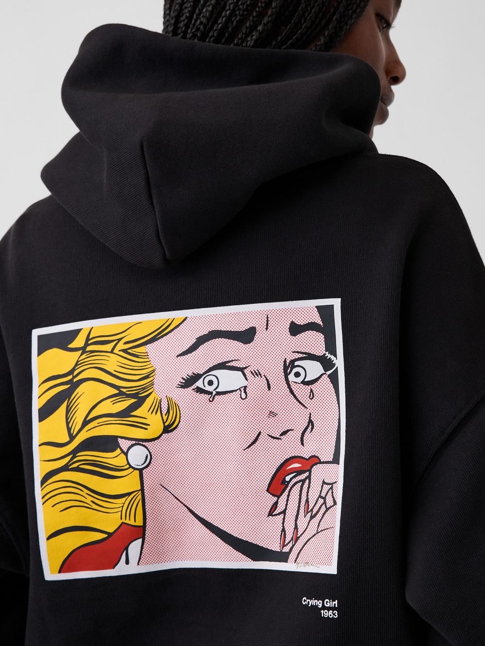 A close-up of a model wearing a black hoodie from the back, featuring a large pop-art graphic print inspired by Roy Lichtenstein's work Crying Girl on the back