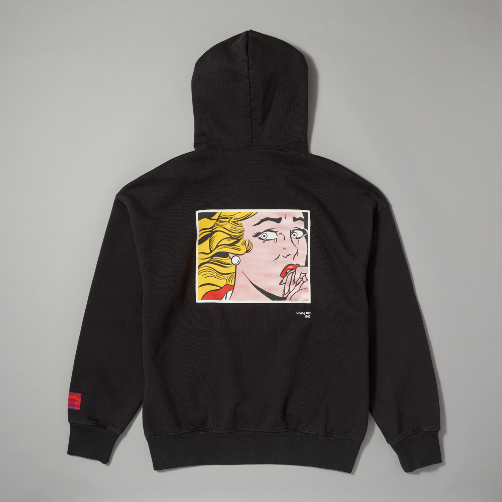 A black pullover hoodie shown from the back, featuring a large square pop-art graphic inspired by Roy Lichtenstein's work Crying Girl on the back. A small red woven label is visible on the left sleeve cuff, set against a solid gray background