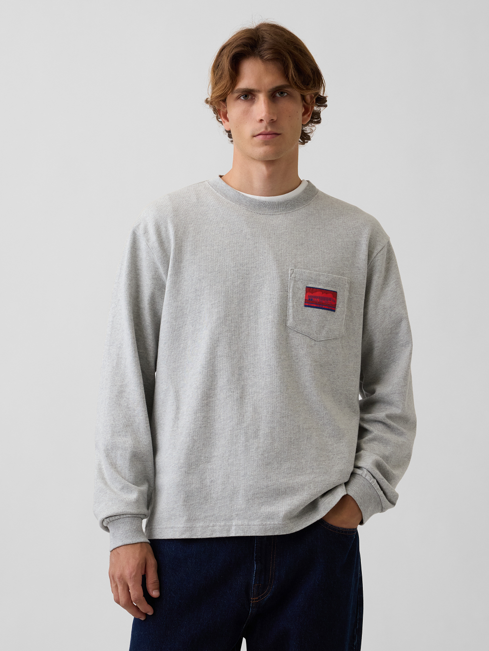 A model wearing a light gray heather crewneck sweatshirt with a chest pocket featuring a red and blue woven label, paired with dark blue jeans, against a plain light gray background