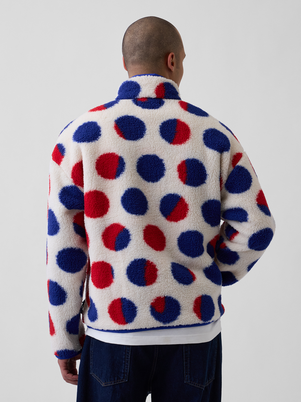 A model viewed from behind wearing a white sherpa fleece jacket with a bold red and blue polka dot pattern inspired by Roy Lichtenstein's work Cathedral #2, paired with dark blue jeans, against a light gray studio background
