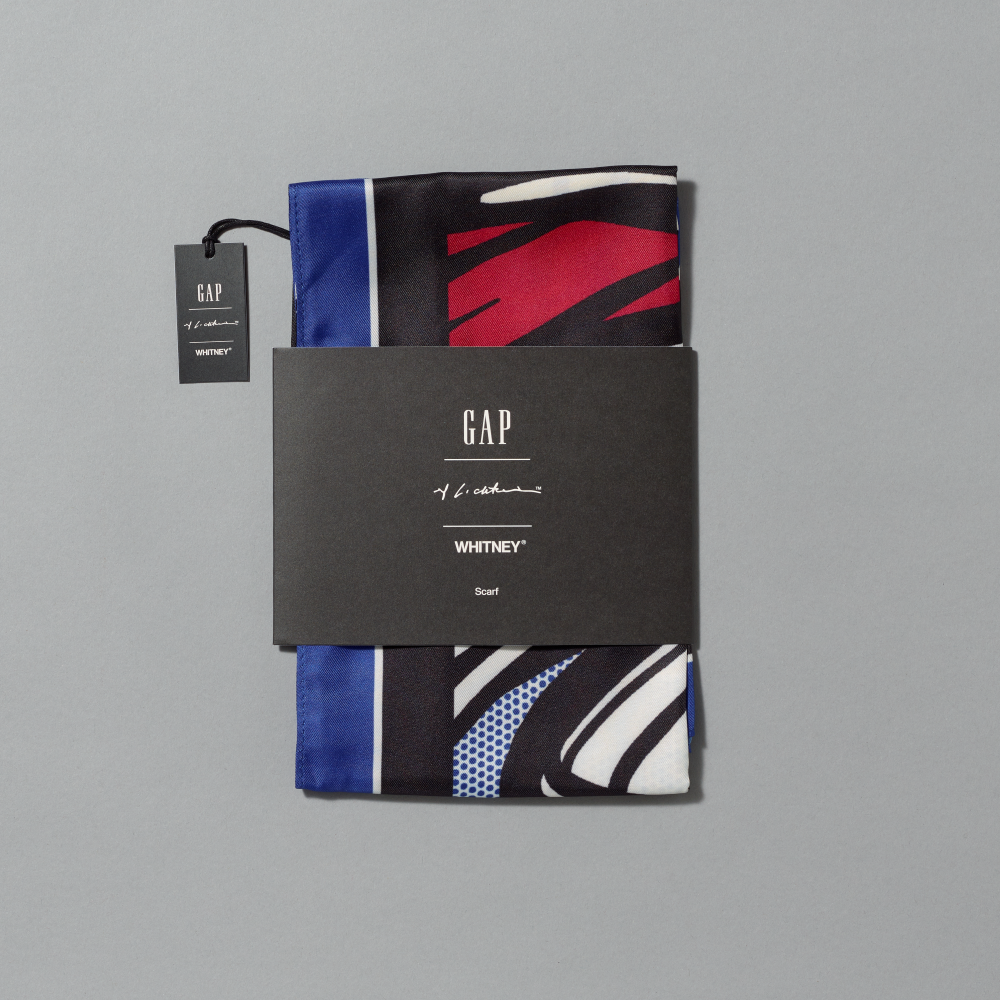 A folded scarf inspired by Roy Lichtenstein's work Little Big Painting wrapped in a black paper band featuring GAP, Lichtenstein and Whitney logos set against a solid gray background