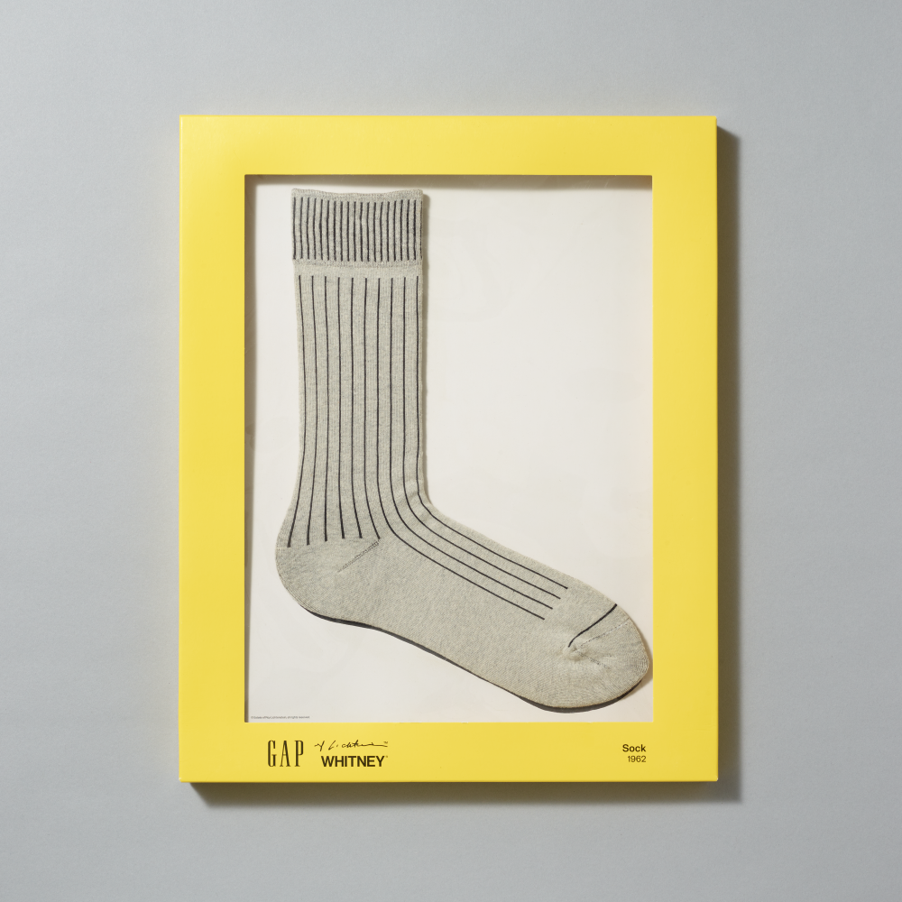 A pair of heather-gray crew socks with thin dark vertical stripes, displayed inside a bright yellow matte box on a light gray background. The frame includes branding for GAP, Lichtenstein and Whitney at the bottom