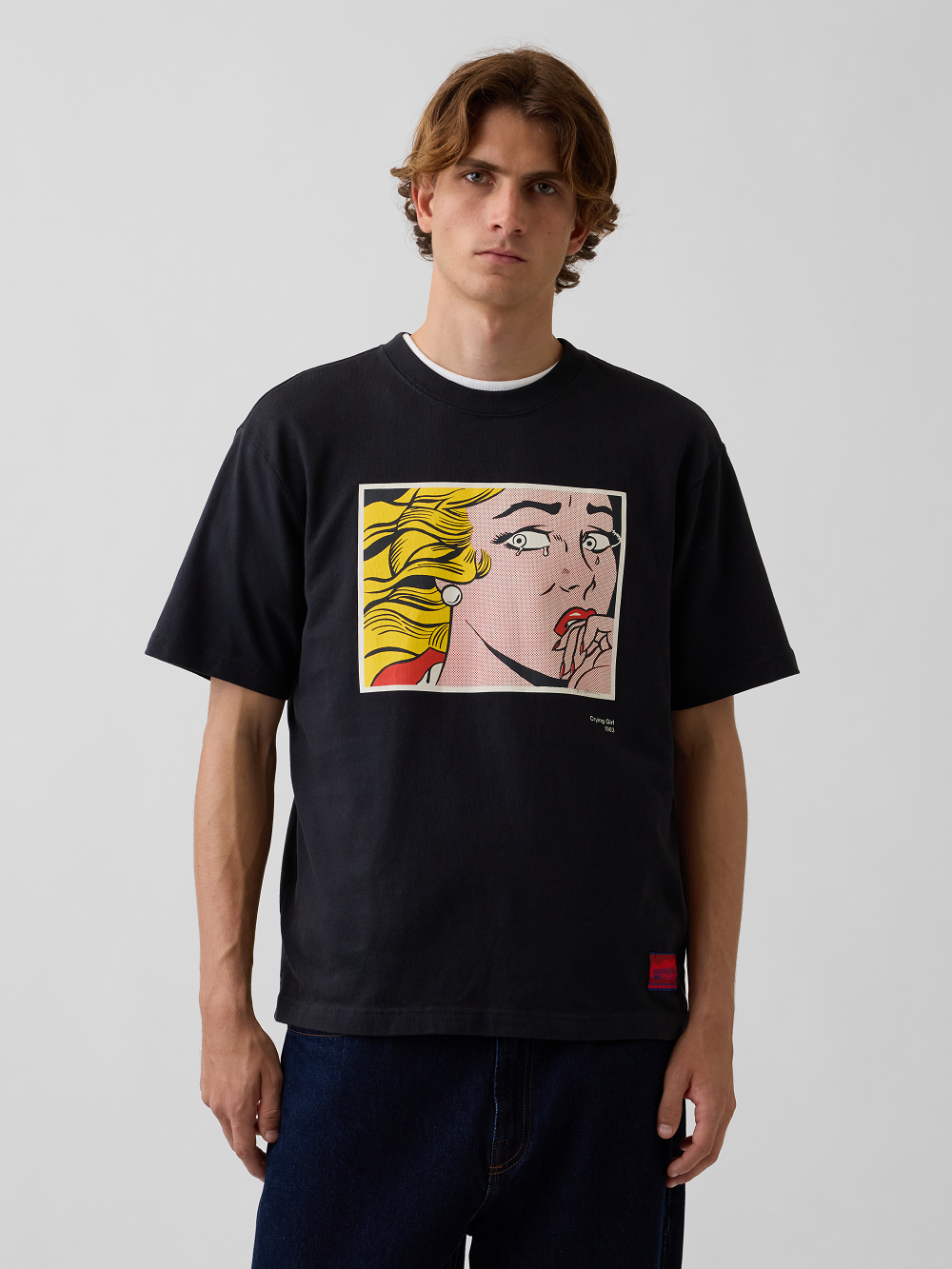 A model wearing a black crewneck t-shirt with a pop-art graphic print inspired by Roy Lichtenstein's work Crying Girl paired with dark blue jeans, against a light gray studio background