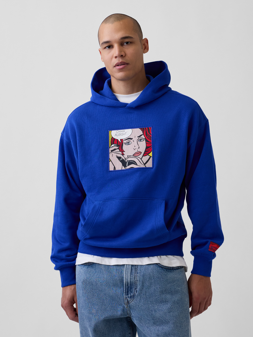 A model wearing a royal blue hoodie with a pop-art graphic print inspired by Roy Lichtenstein's work Ohhh... Alright... on the chest, styled over a white t-shirt and blue jeans, against a plain light gray background