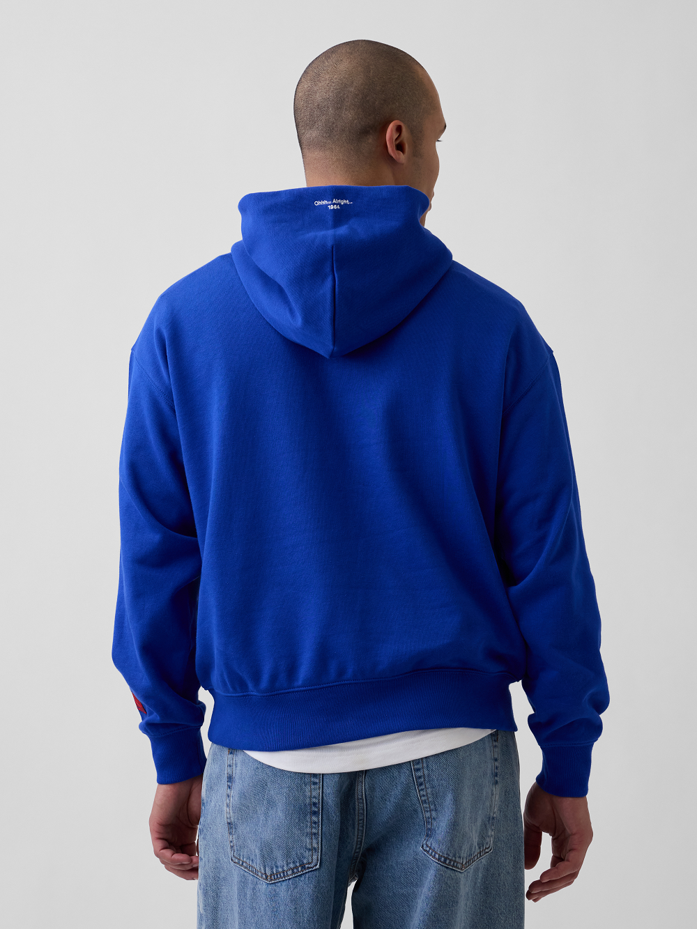 A model viewed from behind wearing a bright royal blue pullover hoodie, a white undershirt, and light-wash blue jeans against a plain light gray studio background