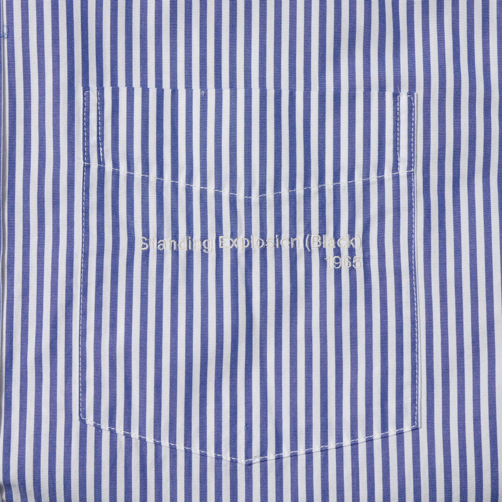Close-up of a blue and white vertically striped pocket on a shirt, with white embroidered text reading 'Standing Explosion (Black) 1965'