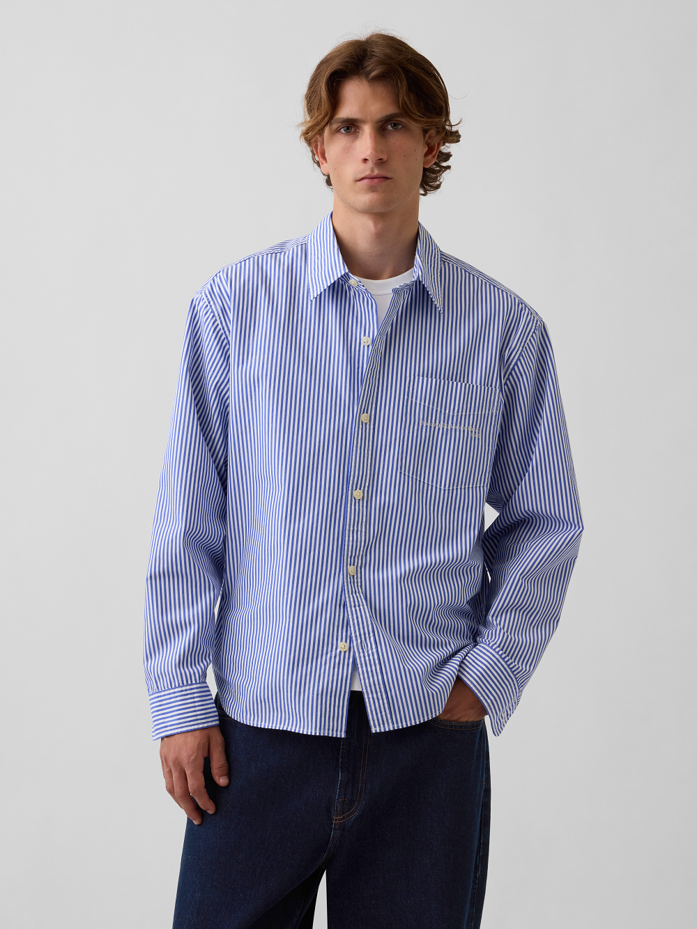 A model wearing a light blue and white vertically striped long-sleeve button-down shirt with a chest pocket, paired with dark blue jeans, against a plain light gray background