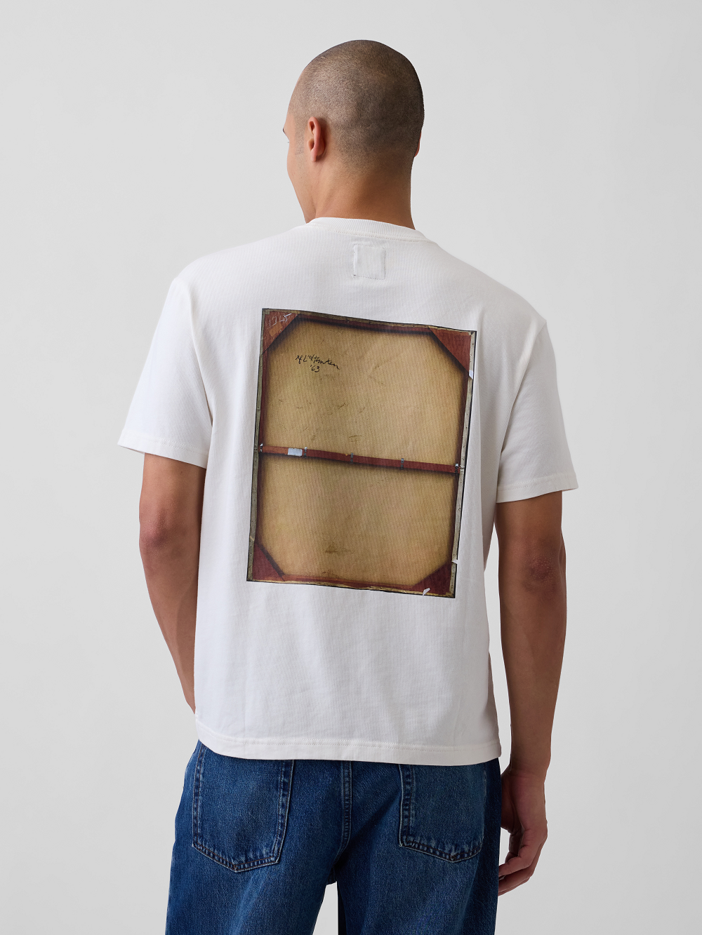 A model viewed from behind wearing a white graphic t-shirt inspired by Roy Lichtenstein's work Girl in Window on the back, paired with blue jeans, against a plain light gray studio background