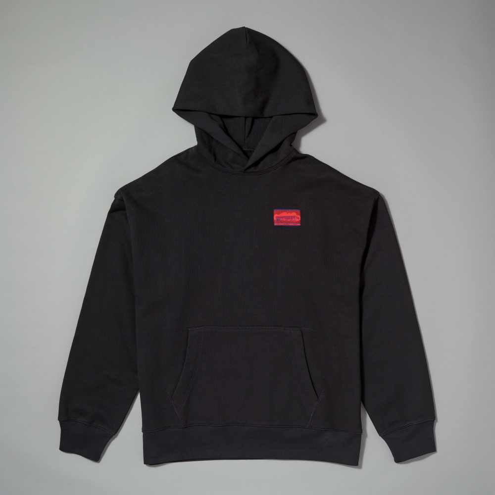 A black pullover hoodie with a small red and blue woven label, laid flat against a neutral gray background