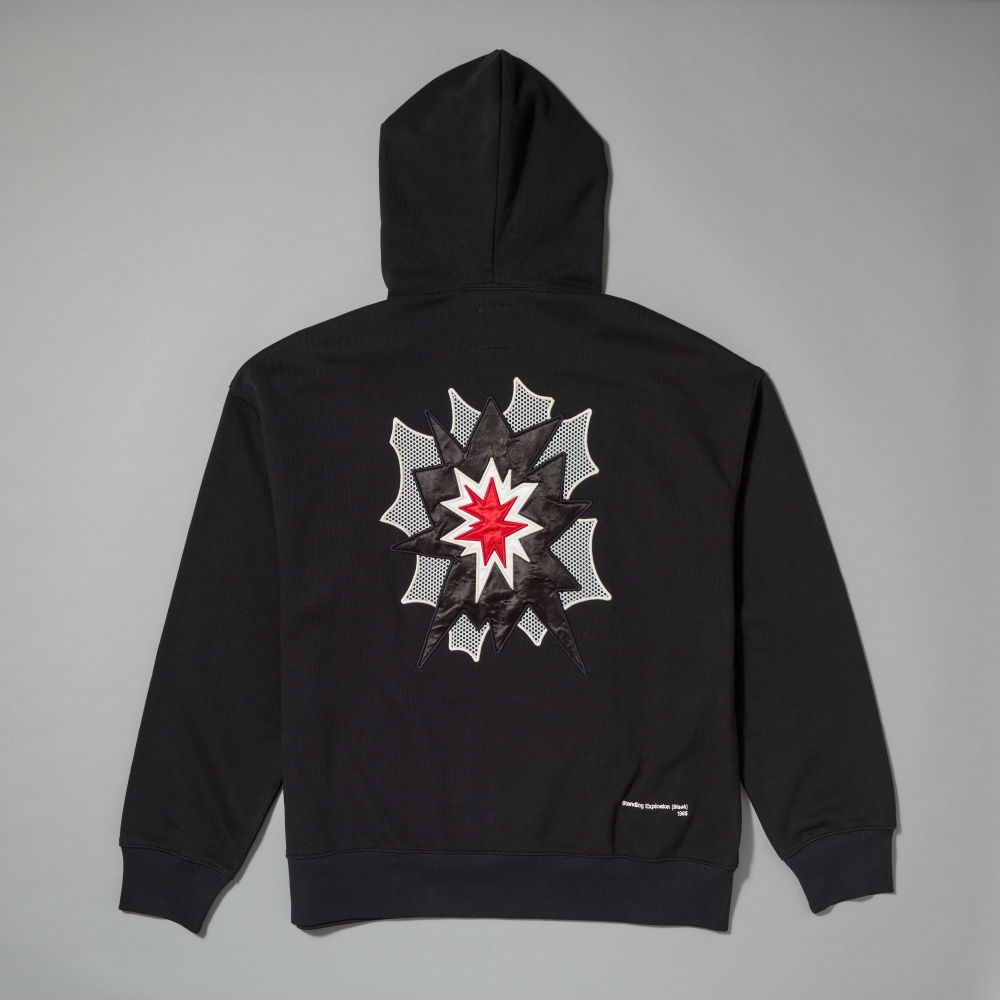 A black pullover hoodie shown from the back, featuring a large graphic print inspired by Roy Lichtenstein's work Standing Explosion in the center. The hoodie is photographed flat against a neutral gray background
