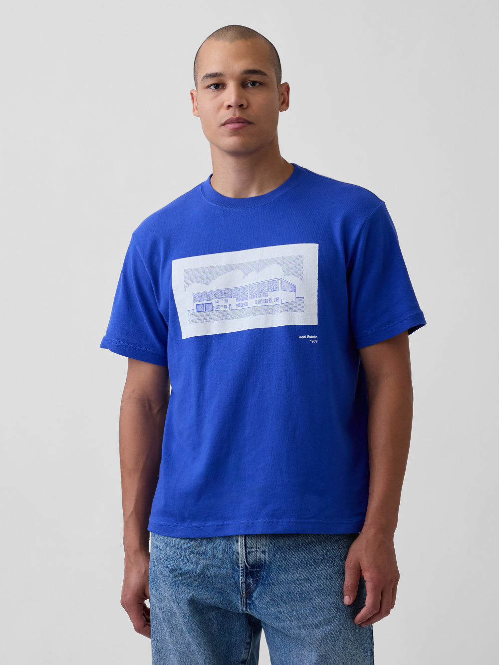 A model wearing a bright royal blue t-shirt with a graphic inspired by Roy Lichtenstein's work Real Estate paired with blue jeans, against a light gray studio background
