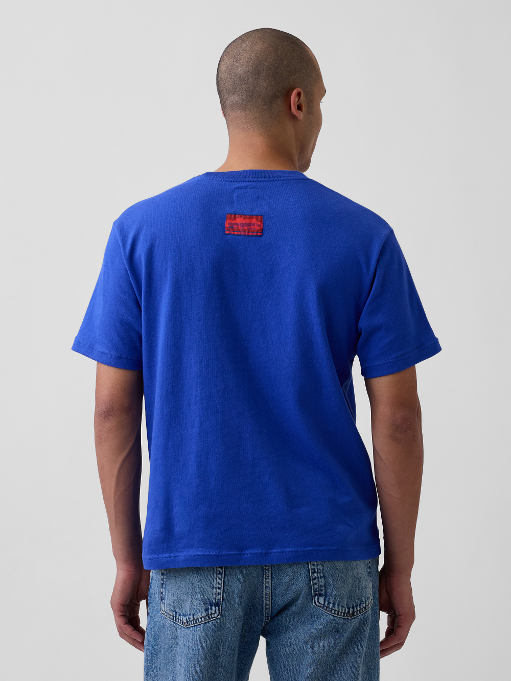 A model viewed from behind wearing a bright royal blue t-shirt with a small red woven label on the upper back, paired with blue jeans, against a light gray studio background