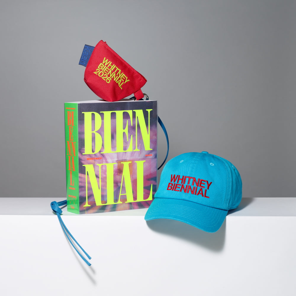Group shot of the Whitney Biennial 2026 exhibition catalogue, red mini lanyard pouch with Whitney Biennial 2026 in neon yellow text, and blue baseball cap with Whitney Biennial in red text on a gray background