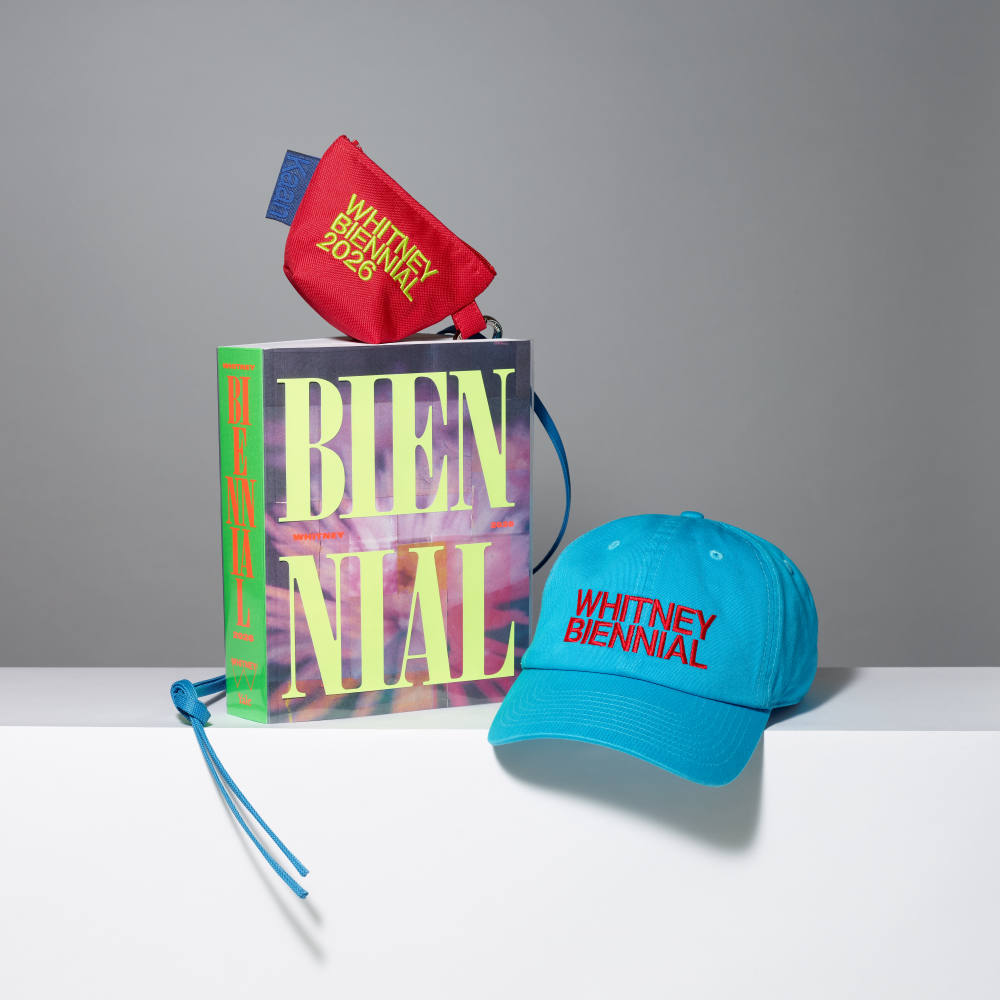 Group shot of the Whitney Biennial 2026 exhibition catalogue, red mini lanyard pouch with Whitney Biennial 2026 in neon yellow text, and blue baseball cap with Whitney Biennial in red text on a gray background