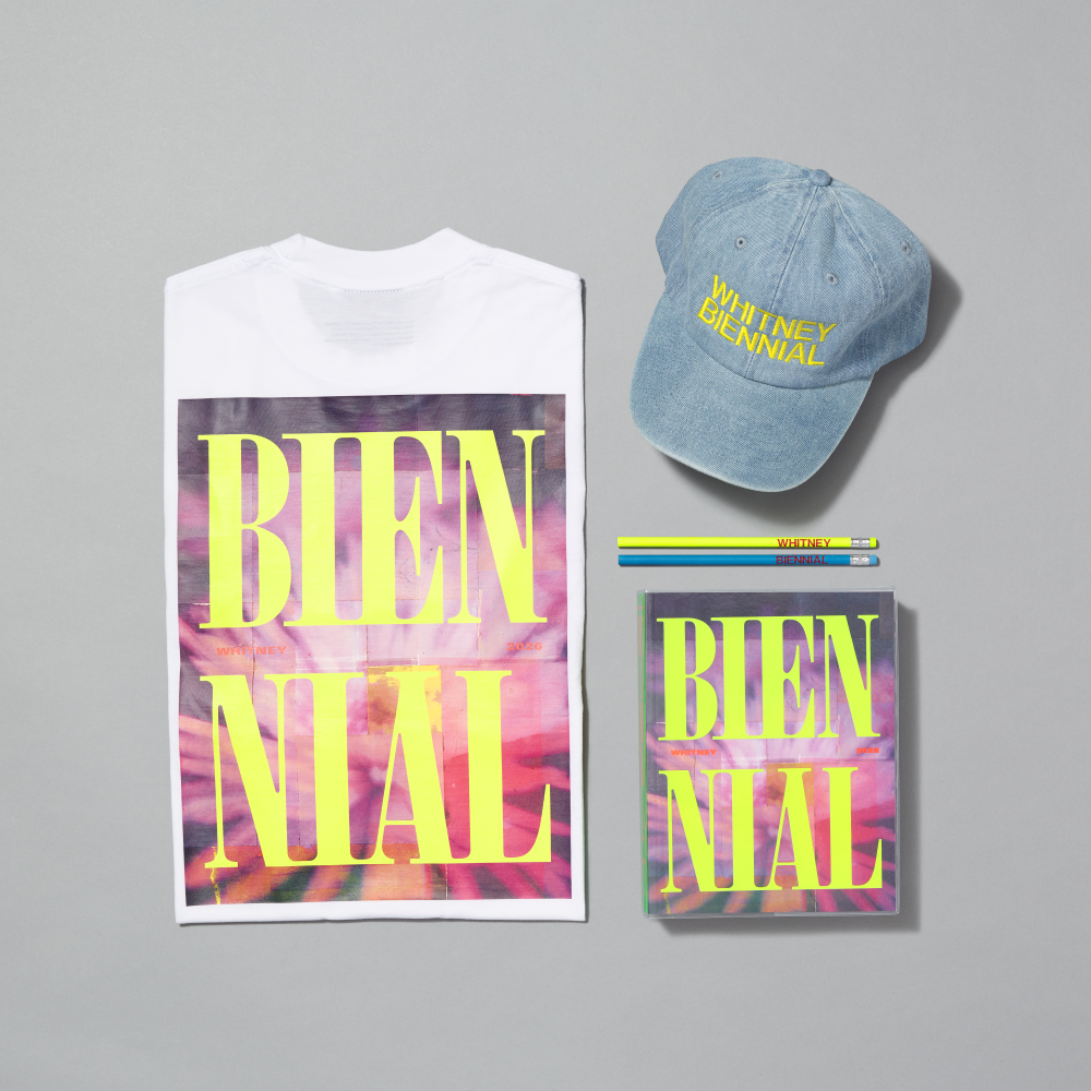 A flat-lay product presentation featuring a white graphic T-shirt, a light blue denim baseball cap, the exhibition catalogue, and two pencils on a solid gray background. The T-shirt and catalogue feature a colorful abstract background with large neon yellow text reading 'BIENNIAL'.