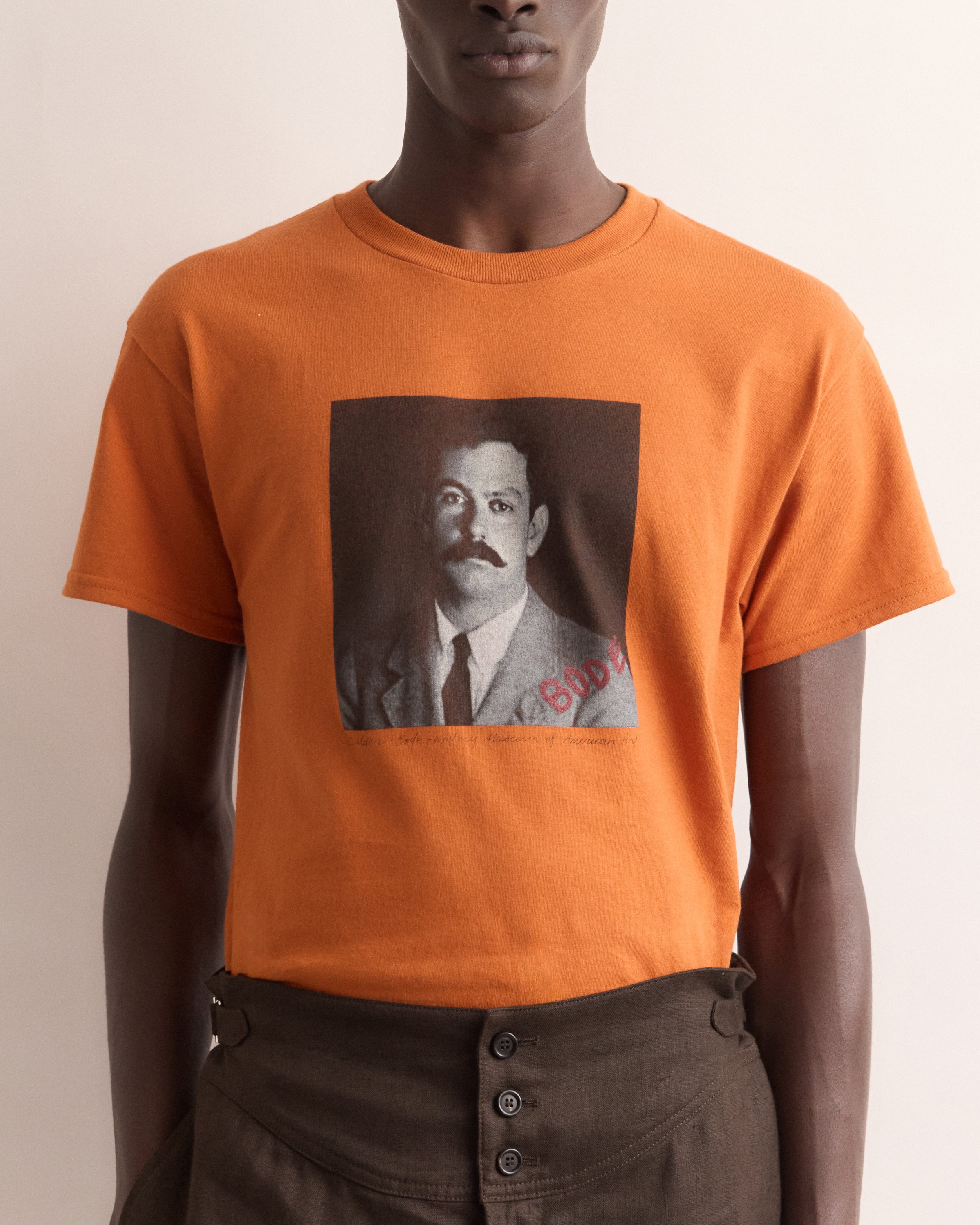 Person wearing an orange t-shirt with a graphic of a man with a mustache and the word 'Buddha'.