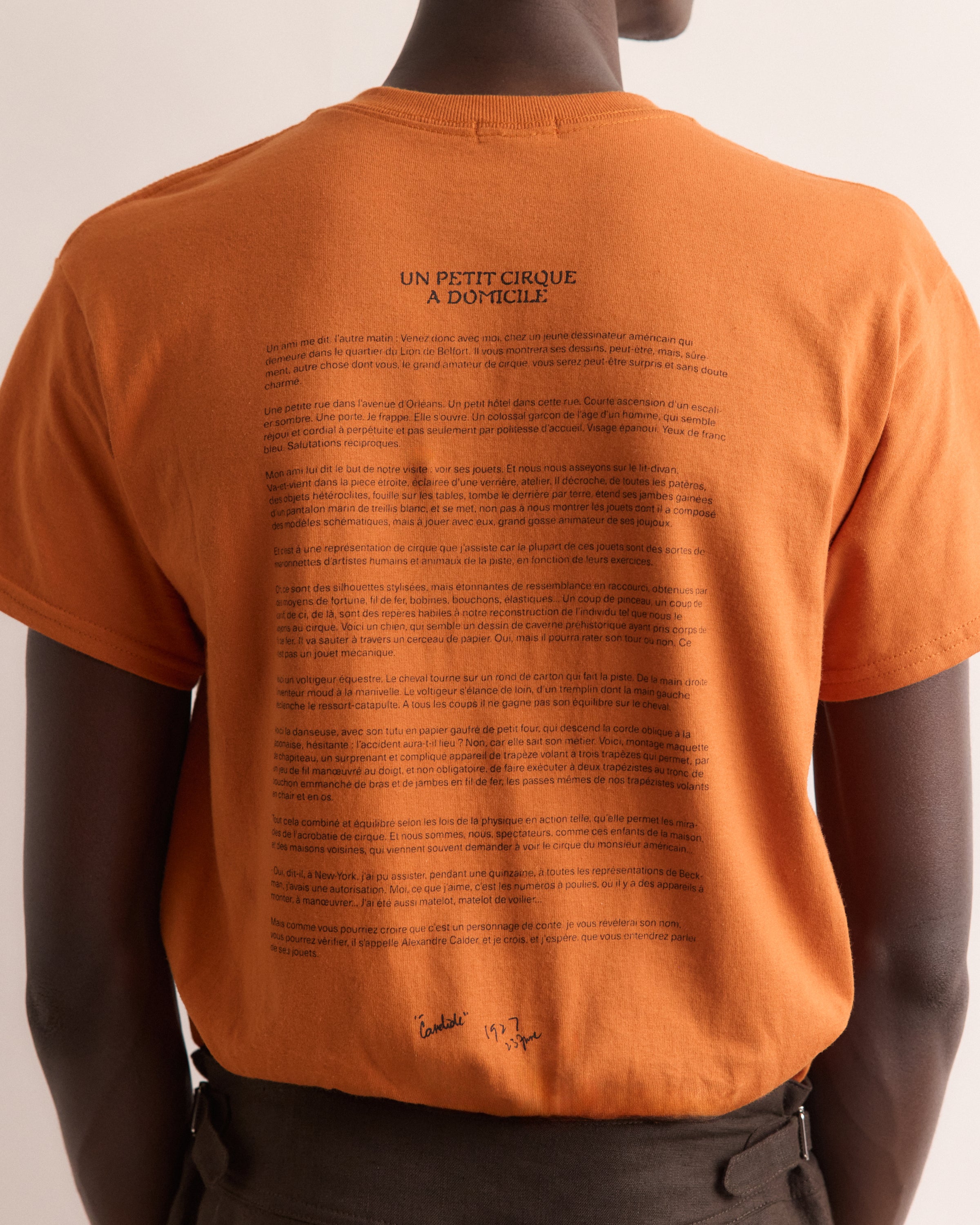 Orange t-shirt with text on the back worn by a person, on a plain background