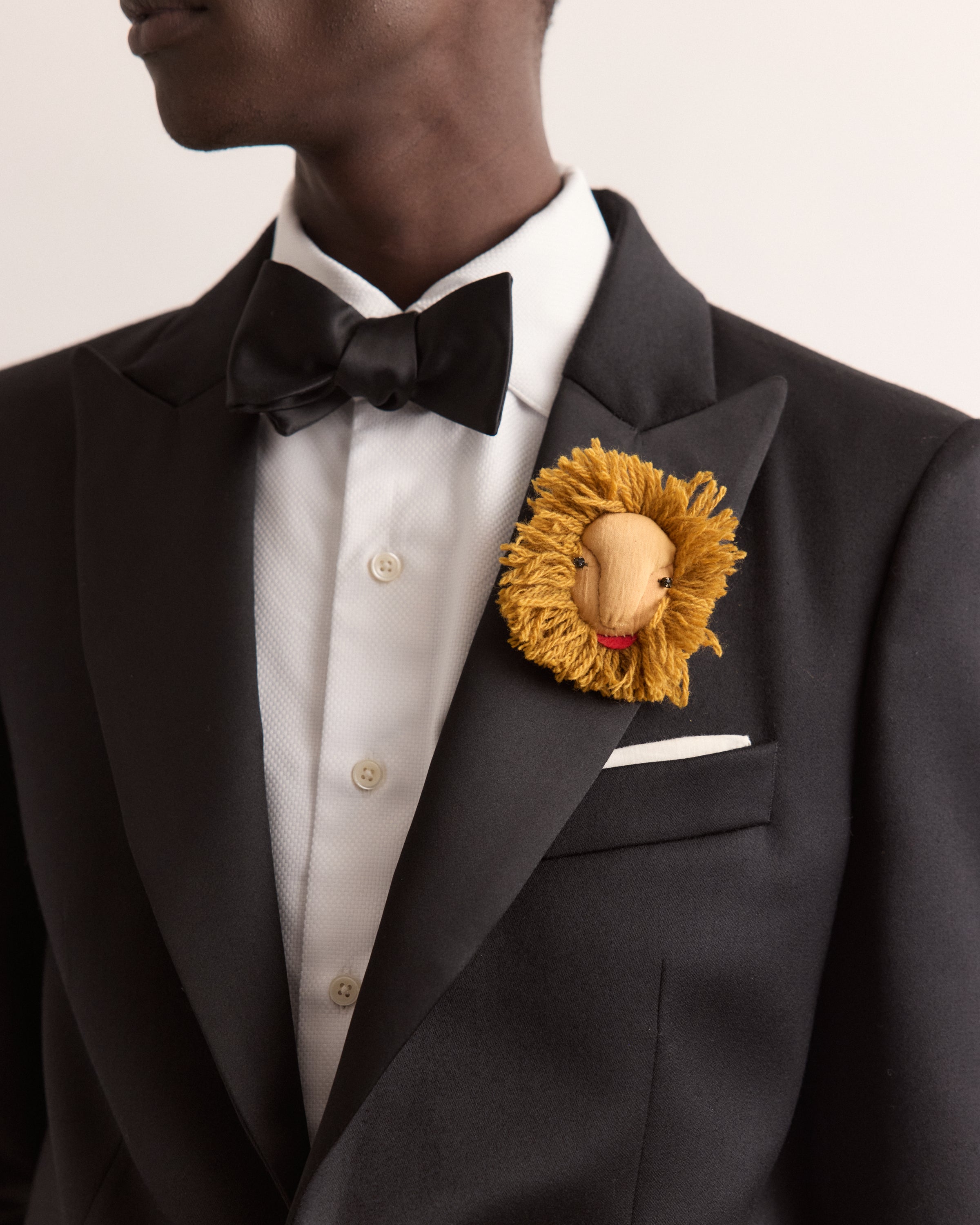 Man wearing a black tuxedo with a lion-themed brooch on a light gray background
